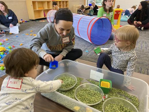 Two young library users and an adult using split peas in a series of bowls for sensory play, while children and caregivers play in the background..