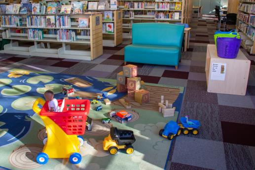 Toys such as play shopping cart, trucks and blocks are spread out in sunshine and picture book shelves can be seen.