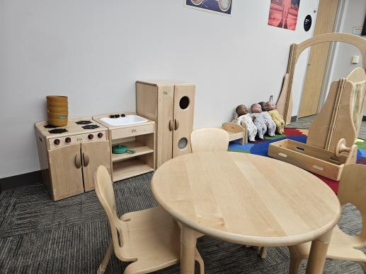 A picture of the play area that includes a play kitchen, play table, baby dolls, play bed, and play steering wheel