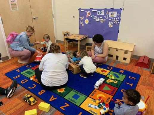 1 adult and child playing with a red truck as well as 2 adults and 3 babies playing with farm animals and a barn on a colorful carpet.