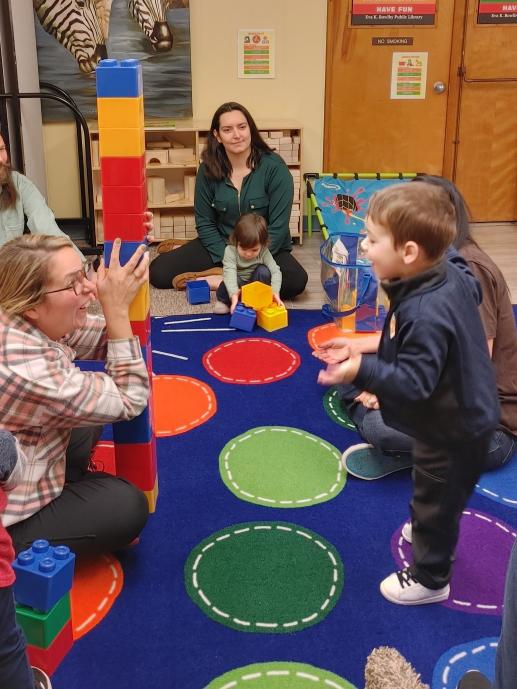 Block Play, mommy and 2yo are equally excited by their tall tower. In background a younger toddler and mommy move blocks around