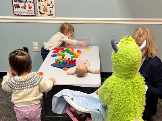Three toddlers are playing with manipulatives on a small table and with baby dolls. One toddler is wearing a monster costume.