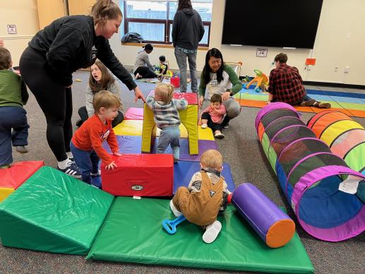 Numerous toddlers and caregivers are interacting. Some of climbing/crawling on floor mats, some are playing with cars and other toys.