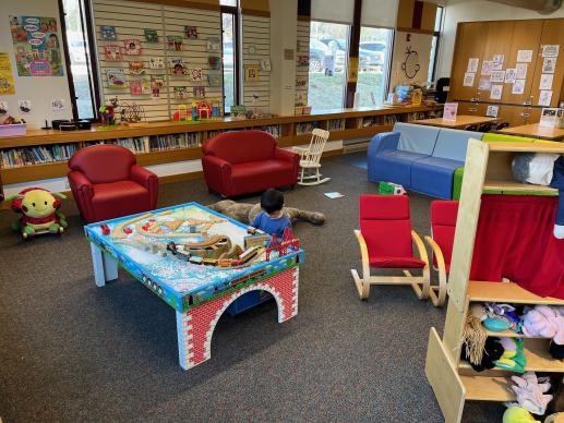 Children's play area with comfortable seating, a train table, puppet theater, toys, board books, puzzles, and activity tables.