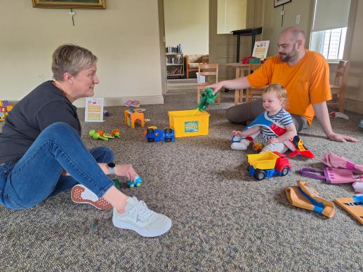 two adults sitting on the ground with a baby, surrounded by toys