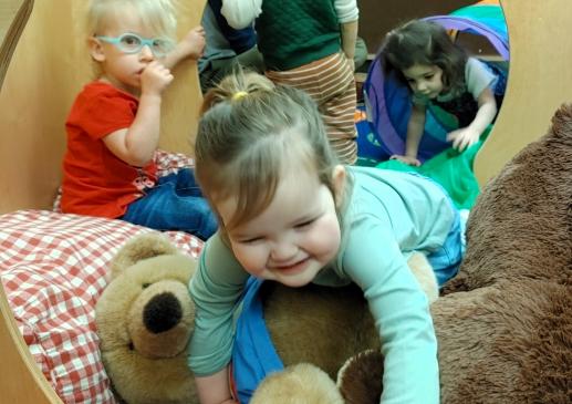 Close up of a smiling child crawling over two large teddy bears in a playhouse. Behind her two children watch and a fourth child crawls through a rainbow tunnel connected to the playhouse