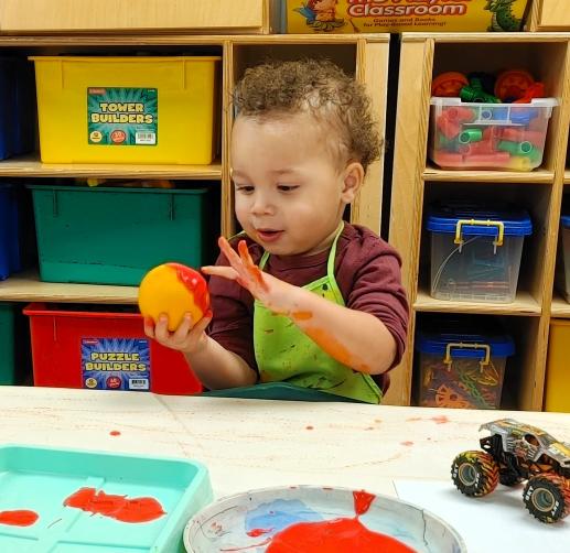 A toddler marvels as he touches the paint on an orange that he is making prints with. Behind him are shelves of various blocks and other manipulative toys.