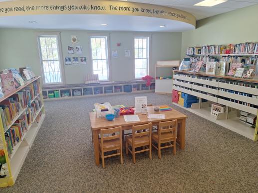 Library children's room with bookshelves, toys stored under a window seat, a child sized wooden table, and a dramatic play center