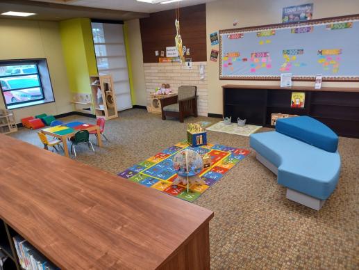 Children's area of library showing colorful rug with toys, blue couch, Lego table, book shelves, and a blue bulletin board.