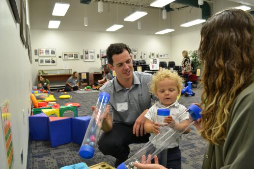 A family playing with sensory tubes with the play room and other families in the background
