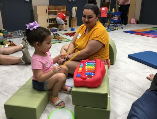 Young girl playing a toy piano with drum sticks with her mother sitting beside her.