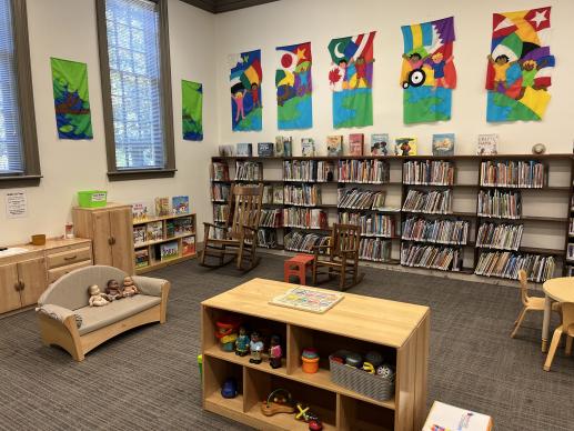 A photo of Larchmont Library's Kid Zone. Colorful banners hang above shelves full of picture books. Wooden shelves full of children's toys can be seen in the foreground, with child sized furniture around the sides of the space.