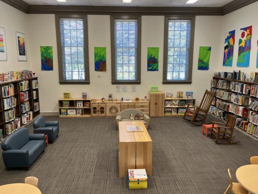 A photo of Larchmont Library's Kid Zone. The sides of the room contain shelves full of picture books. Wooden shelves full of children's toys are in the center of the room, with child sized furniture around the sides of the space and a play kitchen under the windows on the back wall.