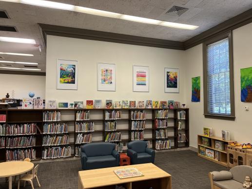 A photo of Larchmont Library's Kid Zone. Colorful posters hang above shelves full of picture books. Child sized furniture and seating surrounds shelves of toys.