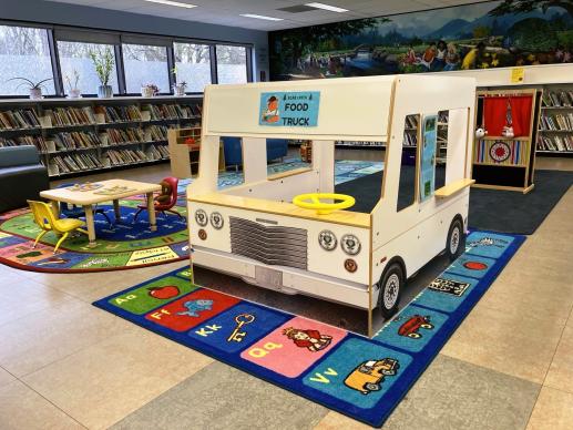 Play food truck on bright carpet in children's area of the library with other toys and furniture in the background.