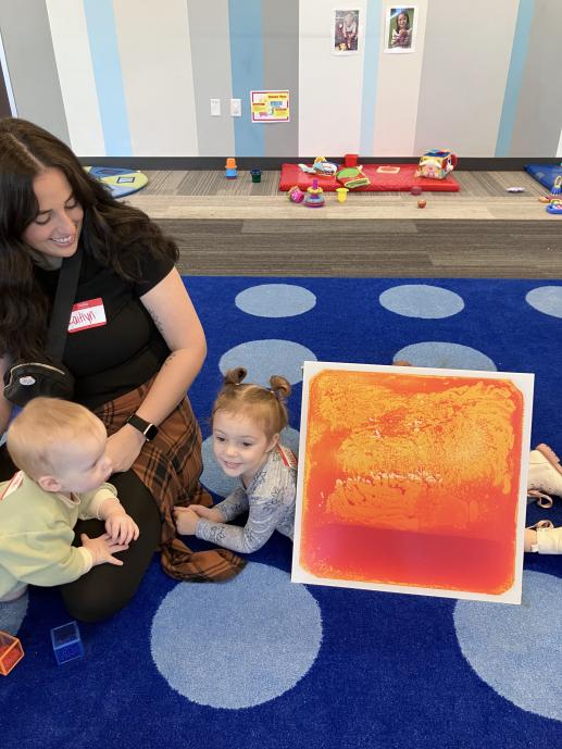A photo of a family with two young children playing with a sensory panel. A toy area is visible in the background.