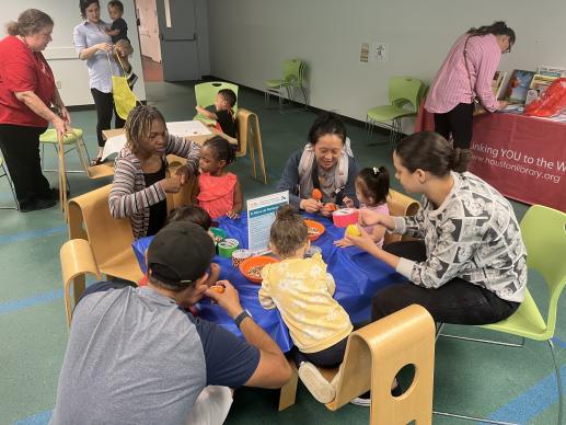 Group of parents and children sitting at small table, working on a craft, while a staff member stands by.