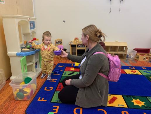 Mother is kneeling on the floor beside her 2 year old daughter playing cooking at a pretend kitchen set. The little girl is holding a red spoon and handing her mom a purple bowl.