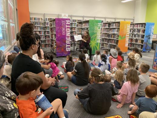 A group of children and their caregivers sitting on the floor, listening to a librarian read at story time.