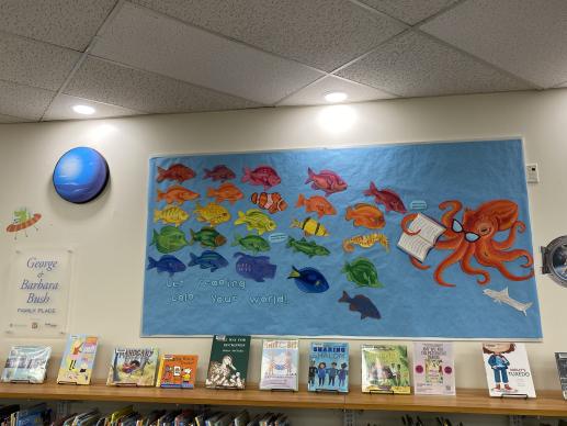 Picture of bulletin board with fish the color of the rainbow swimming toward an octopus with the words: Let reading color your world. To the left of the bulletin board is a foam planet, an alien in a spaceship wall sticker and a sign for the  George & Barbara Bush Family Place. Under the bulletin board are picture books displayed on a shelf