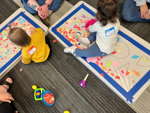 An image showing two toddlers sitting in front of a table set on the floor with shapes drawn on contact paper. They are using small pieces of paper to decorate.