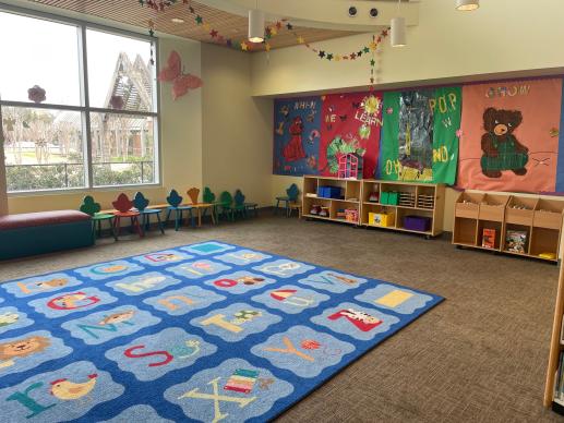 The children's section at the Stella Link library. There is a large storytime rug in the play area. There's a bench and several small chairs in front of the windows. On the far wall, library staff have made artwork of popular children's book characters (Clifford the Big Red Dog, the Very Hungry Caterpillar, Pete the Cat, and Courderoy). There are toys and board books in cubbies.