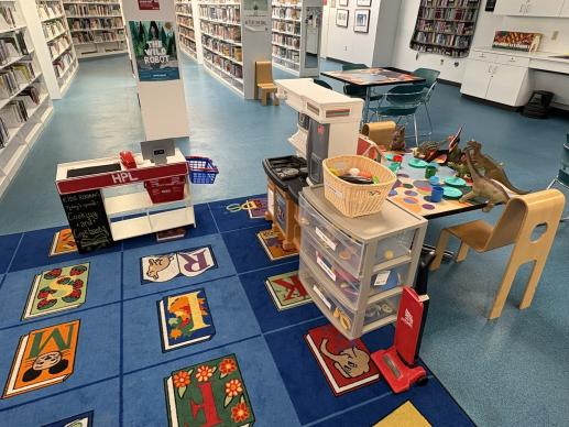 A carpeted children's play area including a play grocery store checkout and play kitchen. A group of toy dinosaurs sits on an adjacent table appearing to have a tea party.