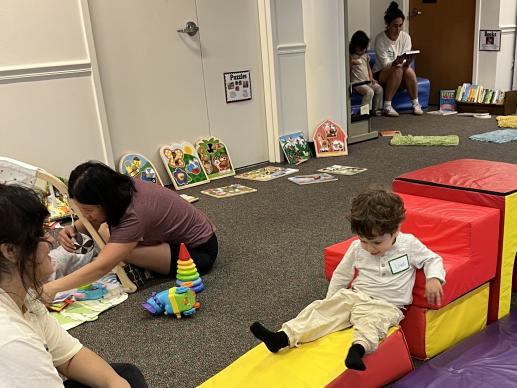 A caregiver plays with a baby while another reads to a young child and a third watches her child slide down a large foam block.