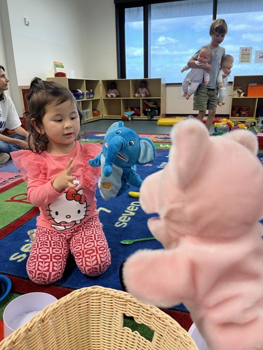 A young girl manipulates a blue elephant hand puppet while the person behind the camera holds up a pig hand puppet.