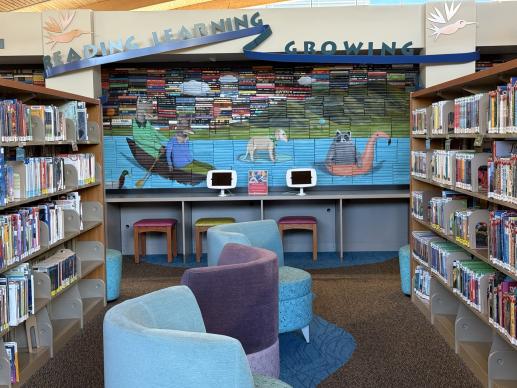 A mural of a lake with local animals painted on the spines of books beside blue and purple furniture and two iPads