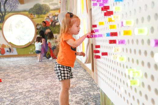 A little girl wearing an orange t-shirt plays with colorful acrylic pegs on the Illumination Station. 