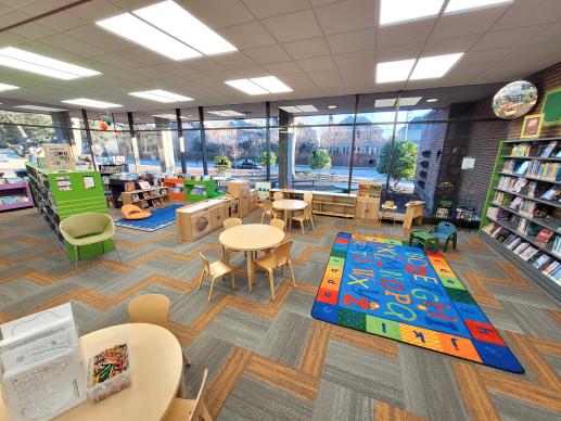 A wide shot of the children's area of Norfolk Public library including activity tables, colorful rugs, different types of seating, a kids zone, and many books. The far corner features ceiling to floor windows.