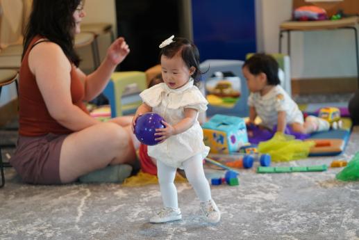 A small girl carries a purple ball while a mother and child play with toys in the background. 