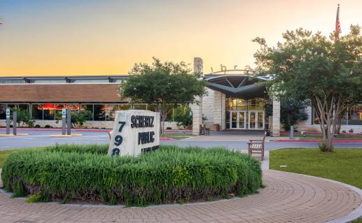 Front exterior of the library building with landscaped grounds and trees, an American flag flying on a flagpole, and a parking lot in the foreground.