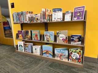 A low wooden bookshelf against a bright yellow wall displaying children’s picture books face-out, with a “Parenting Resources” sign on the top shelf in a library setting.