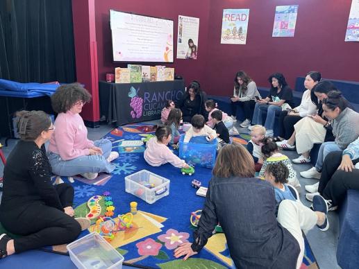 Families and their toddlers are playing on a colored rug. 