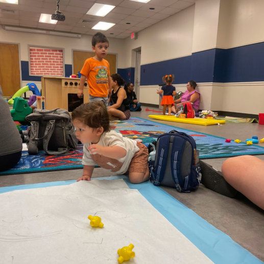 Baby crawling on drawing area, toddler looking around, Resource Professional talking with Parent and toddler.