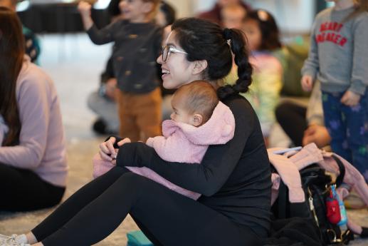 Mother with wearing glasses holds a baby in a pink jacket in her lap in a crowded room. 