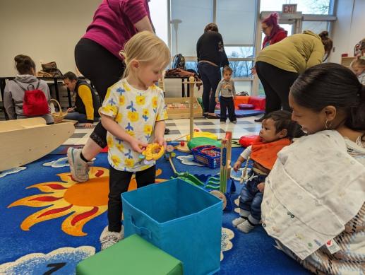 Little girl holding toy looking at mom holding baby on lap. In background see several parents playing with children in various areas