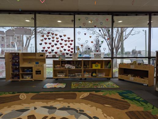 A closer view of a play area in a library. There is a wooden play kitchen, a wooden cabinet full of toys, and a wooden book holder with board books in it.