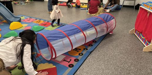 children and caregivers playing; an adult is coaxing a child through a play tunnel in the foreground