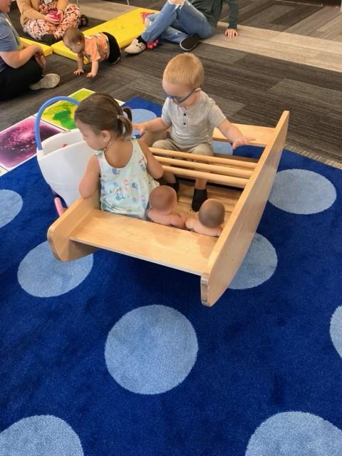 Two toddlers rocking in a wooden boat. One toddler has two baby dolls next to her.