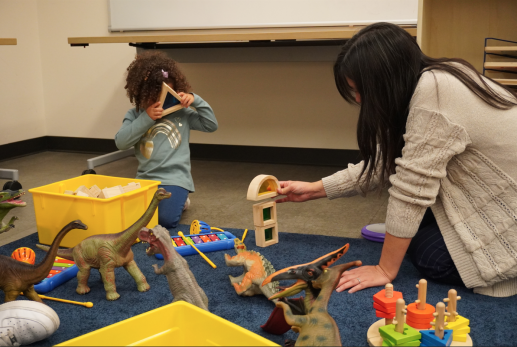 Children's librarian and toddler playing with blocks and dinosaurs on blue carpet.
