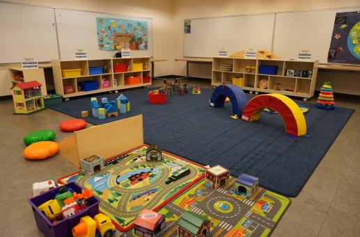 a variety of toys in set up on and around a large blue rug surrounded by shelves full of additional toys in a library meeting room. 