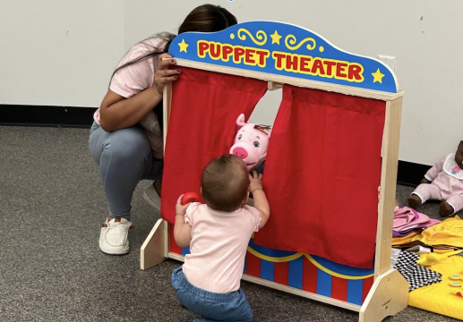 a caregiver reaching behind a puppet theatre curtain to control a puppet a child is playing with
