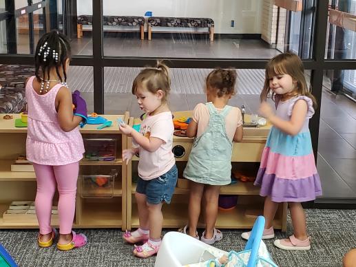 Four young girls are playing together in the toy kitchen area of the library's play space.