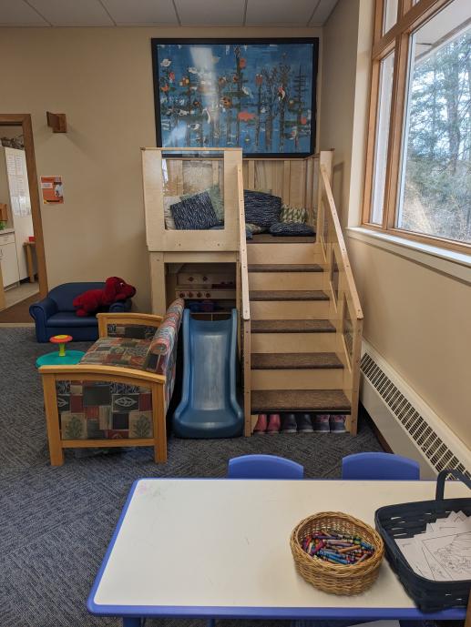 A table in front with crayons and a basket on top. A set of stairs going up to a loft with a slide next to the stairs and a couch next to that. There is a picture on the wall above the loft.