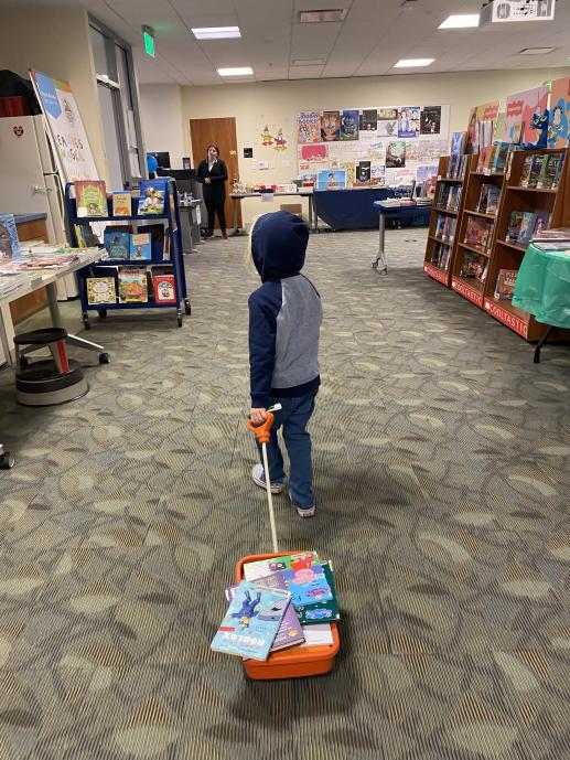 Picture of the back of small boy dragging a miniature orange wagon full of books with adorable and impressive displays of  books  and posters surround him. 